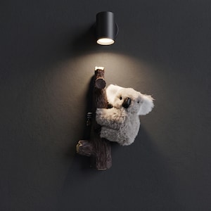 Könnte beinhalten: Eine Wandleuchte mit einem zylindrischen schwarzen Lampenkopf über einer braunen, strukturierten Baumstammskulptur. Eine graue Koala-Plüschfigur mit weißen Innenohren und einer schwarzen Nase ist an der Seite des Baumstamms befestigt.