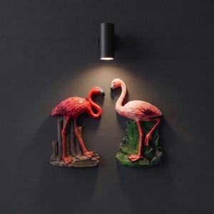 Könnte beinhalten: Zwei an der Wand montierte Flamingoskulpturen in Rosa- und Rottönen. Der linke Flamingo steht auf einem strukturierten braunen Sockel, der rechte auf einem grünen Blättersockel. Beide Figuren werden von einer schwarzen zylindrischen Wandleuchte beleuchtet.