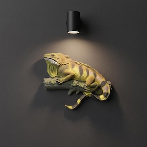 Puede incluir: Una escultura de pared de una iguana posada sobre una rama texturizada. La iguana presenta un cuerpo amarillo y verde con rayas horizontales de color marrón oscuro a lo largo del lomo y la cola. Una pequeña lámpara cilíndrica negra está montada arriba.