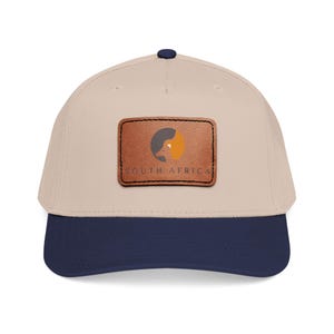 Peut inclure: Casquette de baseball beige avec une visière bleu marine. La casquette présente un écusson en cuir marron avec un graphisme d'oiseau et le texte "SOUTH AFRICA" en noir.