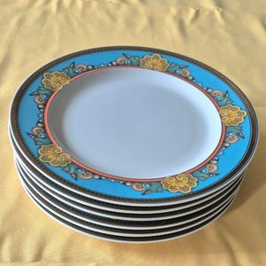 May include: A stack of six white ceramic plates with a light blue border featuring a floral design in yellow, orange, and brown. The plates have a gold and black trim and a white center.
