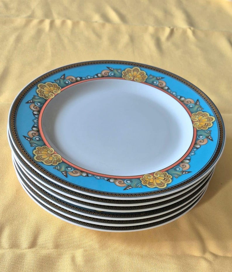 May include: A stack of six white ceramic plates with a light blue border featuring a floral design in yellow, orange, and brown. The plates have a gold and black trim and a white center.