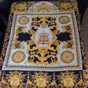 May include: A decorative textile featuring a repeating pattern of gold baroque designs on a navy blue and light blue background. The design includes ornate scrolls, medallions, and a central emblem, creating a luxurious aesthetic. The textile is likely a scarf or a decorative cloth.