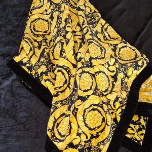 May include: A black and gold patterned scarf with a floral design. The scarf has a black velvet border. The scarf is folded on a black surface.