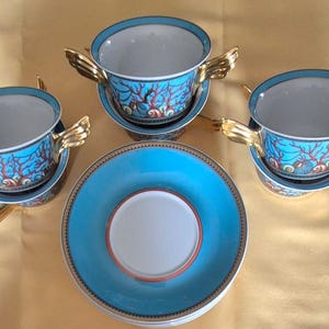 May include: A set of four ceramic teacups and saucers. The cups and saucers are turquoise with a coral reef design. The cups have gold-colored wing-shaped handles. The saucers have a gold border.