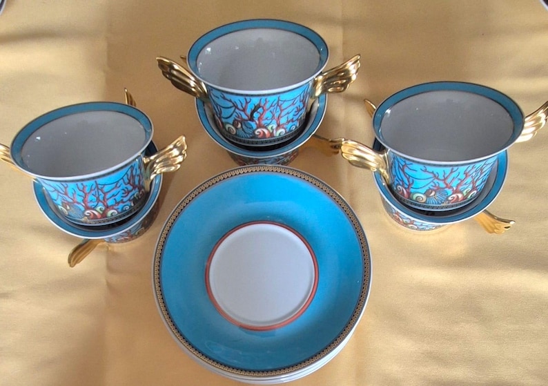 May include: A set of four ceramic teacups and saucers. The cups and saucers are turquoise with a coral reef design. The cups have gold-colored wing-shaped handles. The saucers have a gold border.