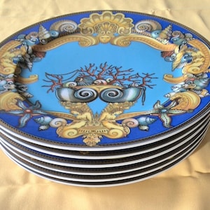 May include: A stack of oval-shaped decorative plates. Each plate features a blue border and a central design with a light blue background. The design includes coral, seashells, and ornate gold accents. The plates have a white interior.