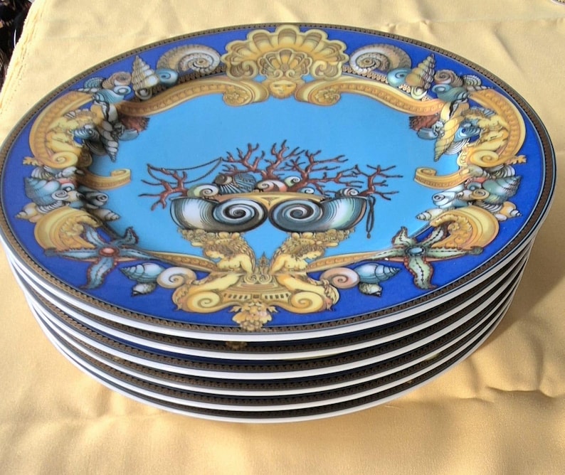 May include: A stack of oval-shaped decorative plates. Each plate features a blue border and a central design with a light blue background. The design includes coral, seashells, and ornate gold accents. The plates have a white interior.