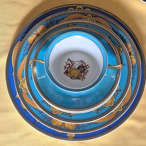 May include: A stack of fine china dinnerware in shades of blue, teal, and gold. The set includes plates of varying sizes, a saucer, and a teacup. The designs feature ornate patterns and a central emblem. The china is arranged on a yellow surface.