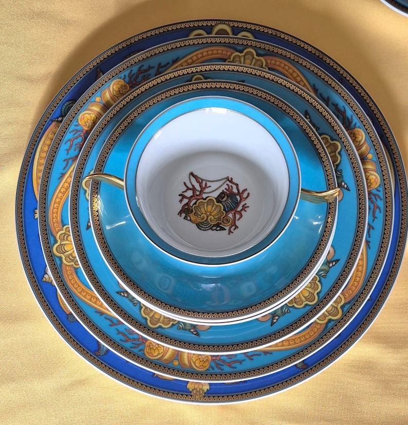 May include: A stack of fine china dinnerware in shades of blue, teal, and gold. The set includes plates of varying sizes, a saucer, and a teacup. The designs feature ornate patterns and a central emblem. The china is arranged on a yellow surface.