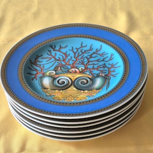 May include: A stack of decorative plates with a blue rim and a central design featuring coral and seashells. The plates have a gold and black border and a light blue center. The plates are stacked on top of each other.