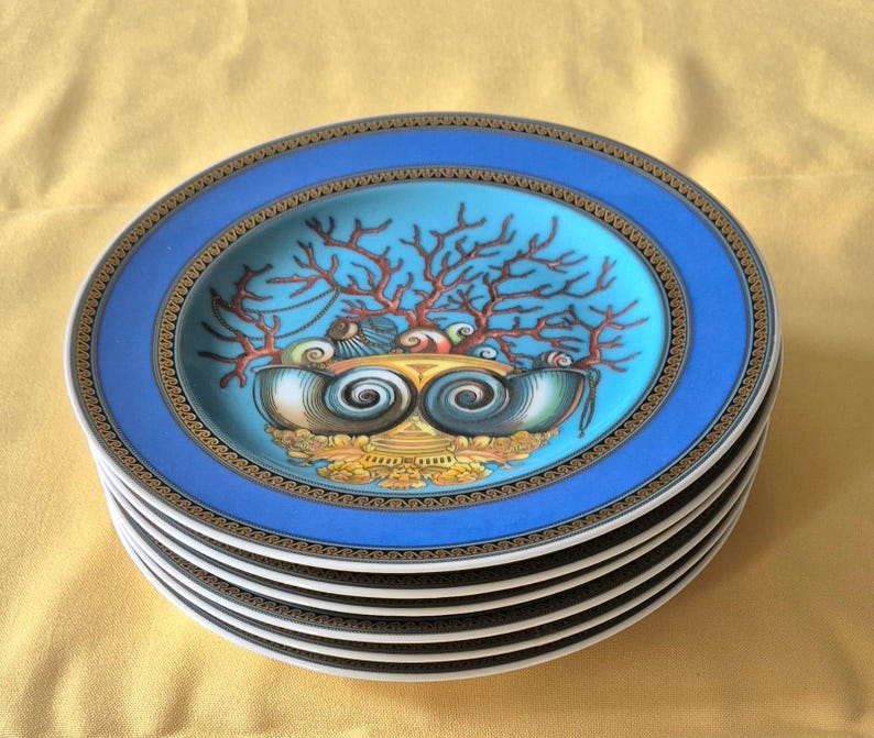 May include: A stack of decorative plates with a blue rim and a central design featuring coral and seashells. The plates have a gold and black border and a light blue center. The plates are stacked on top of each other.