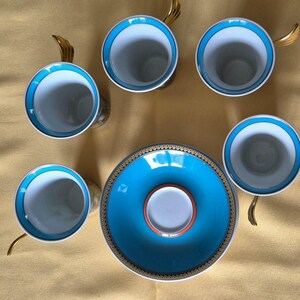 May include: Overhead view of a porcelain tea set with six cups and saucers. The cups have a white interior, a turquoise blue rim, and gold-colored handles. The saucer is turquoise blue with a gold and black border.