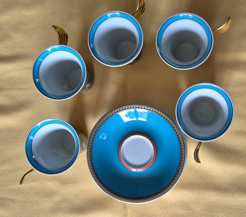 May include: Overhead view of a porcelain tea set with six cups and saucers. The cups have a white interior, a turquoise blue rim, and gold-colored handles. The saucer is turquoise blue with a gold and black border.