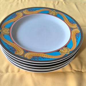 May include: A stack of oval-shaped ceramic plates. Each plate features a white center, a turquoise border with gold and orange decorative elements, and a dark brown outer rim. The plates are stacked on a yellow surface.