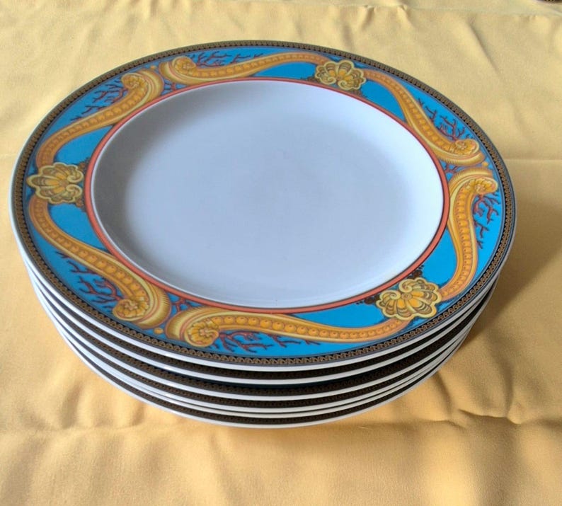 May include: A stack of oval-shaped ceramic plates. Each plate features a white center, a turquoise border with gold and orange decorative elements, and a dark brown outer rim. The plates are stacked on a yellow surface.