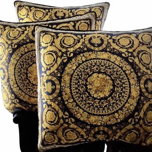 May include: Four decorative throw pillows with a black and gold baroque design. The pillows feature a repeating circular pattern with floral and scroll motifs. The gold elements contrast against the dark background, creating a luxurious aesthetic.