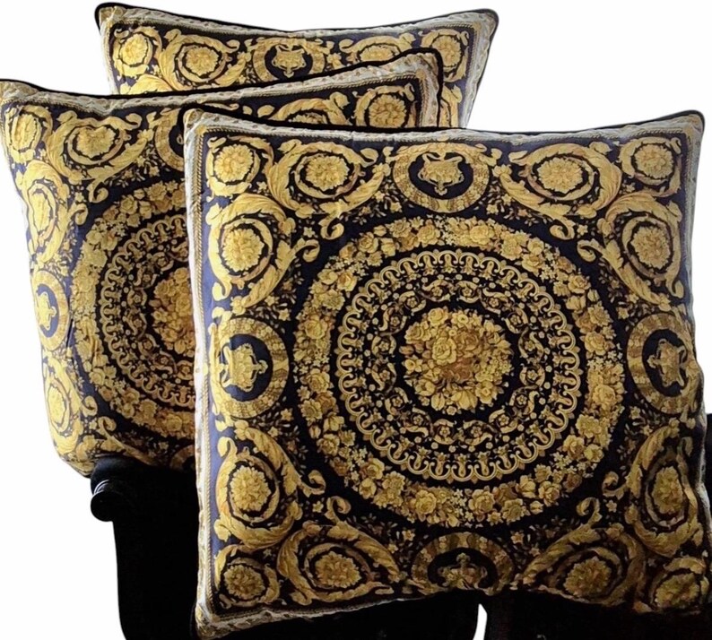 May include: Four decorative throw pillows with a black and gold baroque design. The pillows feature a repeating circular pattern with floral and scroll motifs. The gold elements contrast against the dark background, creating a luxurious aesthetic.