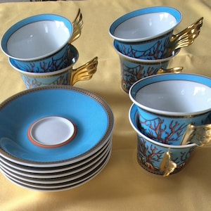 May include: A set of six teacups and saucers. The cups are light blue with a coral design and gold wing-shaped handles. The saucers are light blue with a gold rim and a white center. The teacups and saucers are stacked on a yellow surface.