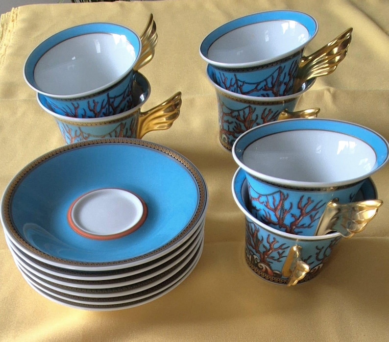 May include: A set of six teacups and saucers. The cups are light blue with a coral design and gold wing-shaped handles. The saucers are light blue with a gold rim and a white center. The teacups and saucers are stacked on a yellow surface.