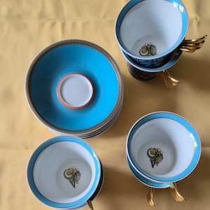 May include: A set of porcelain teacups and saucers. The cups and saucers are white with a turquoise blue border and a gold trim. The cups have a gold wing-shaped handle and a seashell design inside. The saucers are stacked.