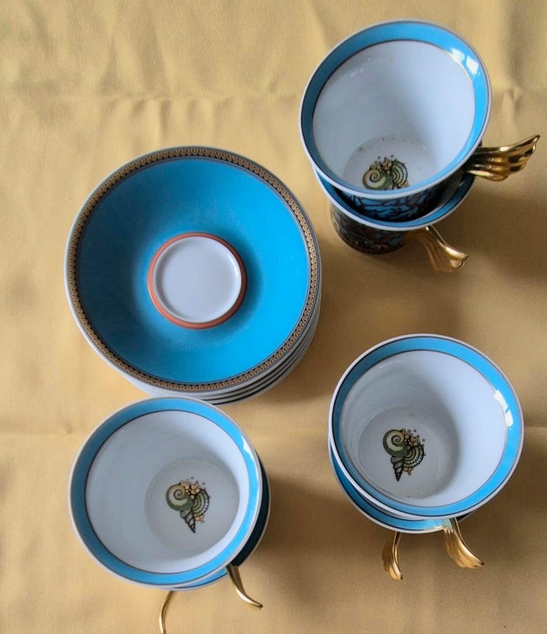 May include: A set of porcelain teacups and saucers. The cups and saucers are white with a turquoise blue border and a gold trim. The cups have a gold wing-shaped handle and a seashell design inside. The saucers are stacked.