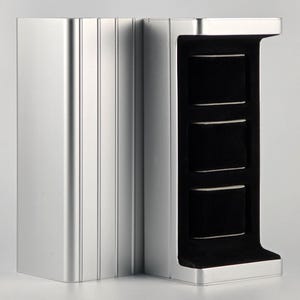 Könnte beinhalten: Ein silberfarbenes Schmuck-Display mit modernem Design. Das Gehäuse hat eine vertikale, rechteckige Form mit drei schwarzen, samtgefütterten Fächern zur Aufbewahrung von Schmuck. Die Außenseite hat eine gerippte Textur auf einer Seite.