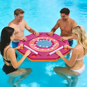 May include: A floating octagonal game table in magenta and orange with a central blue circular area featuring white tiles with black markings. The table includes integrated cup holders and slots for tile racks, designed for use in a swimming pool.