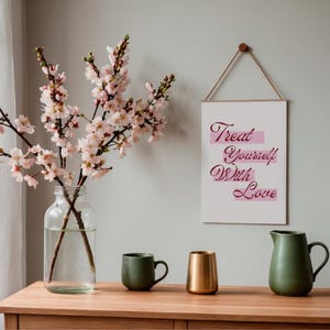 K&ouml;nnte beinhalten: Auf einem Holztisch stehen eine klare Glasvase mit rosa bl&uuml;henden Zweigen, eine gr&uuml;ne Tasse, eine goldfarbene Vase und ein gr&uuml;ner Krug. Ein wei&szlig;es Schild mit dem Text "Treat Yourself With Love" h&auml;ngt an der Wand.