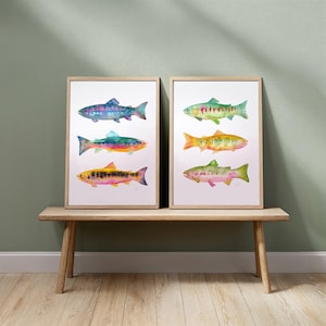 May include: Two framed art prints featuring watercolor paintings of colorful fish. Each print displays three fish in vibrant hues of blue, green, yellow, and pink. The artwork is displayed on a wooden bench against a muted green wall.