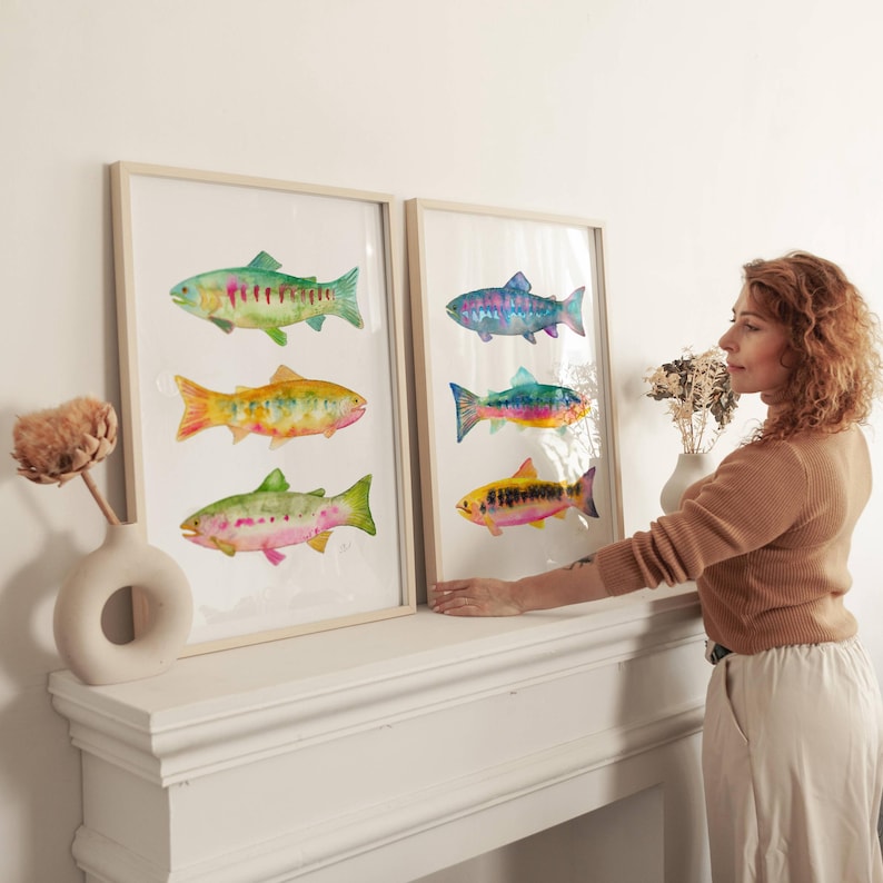 May include: Two framed watercolor prints featuring colorful fish illustrations. Each print showcases three different fish, with vibrant hues of green, yellow, pink, and blue. The frames are light-colored, and the artwork is displayed on a white surface.