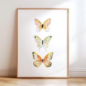Peut inclure: Impression encadr&eacute;e avec trois illustrations de papillons &agrave; l'aquarelle. Les papillons ont des ailes d&eacute;licates dans les tons de rose, jaune et orange, avec de subtiles marques noires. L'&oelig;uvre est sur fond blanc, encadr&eacute;e de bois clair.
