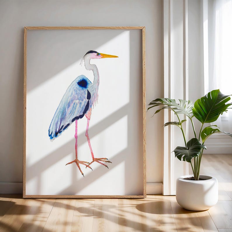 Puede incluir: Pintura a la acuarela de una garza azul en un marco de madera. El ave tiene plumas azules, grises y negras, patas rosas y un pico amarillo. La obra de arte se exhibe en una pared blanca, con una planta en una maceta blanca.