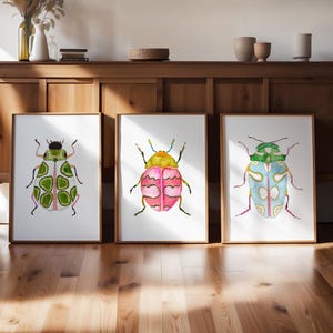 May include: Three framed watercolor paintings of colorful beetles. The beetles are green, pink, and blue with black outlines. The paintings are on a white background and are framed in natural wood.