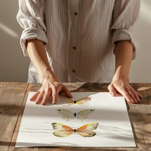 Peut inclure: Impression d'art de papillons &agrave; l'aquarelle repr&eacute;sentant trois papillons dans des tons de jaune, d'orange et de marron. Les papillons sont peints sur un fond blanc. L'&oelig;uvre est expos&eacute;e sur une table en bois.