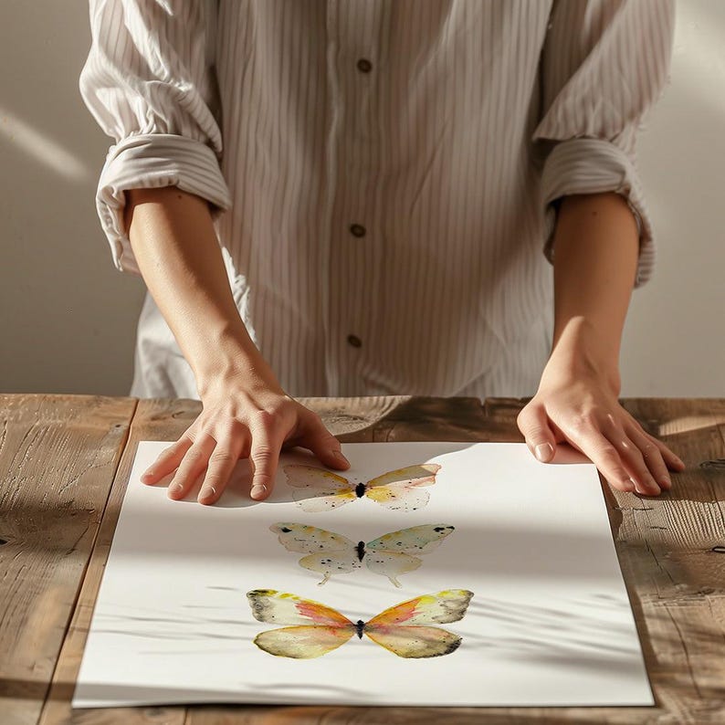 Peut inclure: Impression d'art de papillons &agrave; l'aquarelle repr&eacute;sentant trois papillons dans des tons de jaune, d'orange et de marron. Les papillons sont peints sur un fond blanc. L'&oelig;uvre est expos&eacute;e sur une table en bois.
