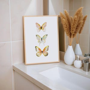 Peut inclure: Une affiche encadr&eacute;e avec trois papillons &agrave; l'aquarelle dans des tons de jaune, rose et vert. L'&oelig;uvre est pr&eacute;sent&eacute;e sur un fond blanc dans un cadre en bois clair. L'affiche est pos&eacute;e sur un comptoir blanc pr&egrave;s d'un lavabo.