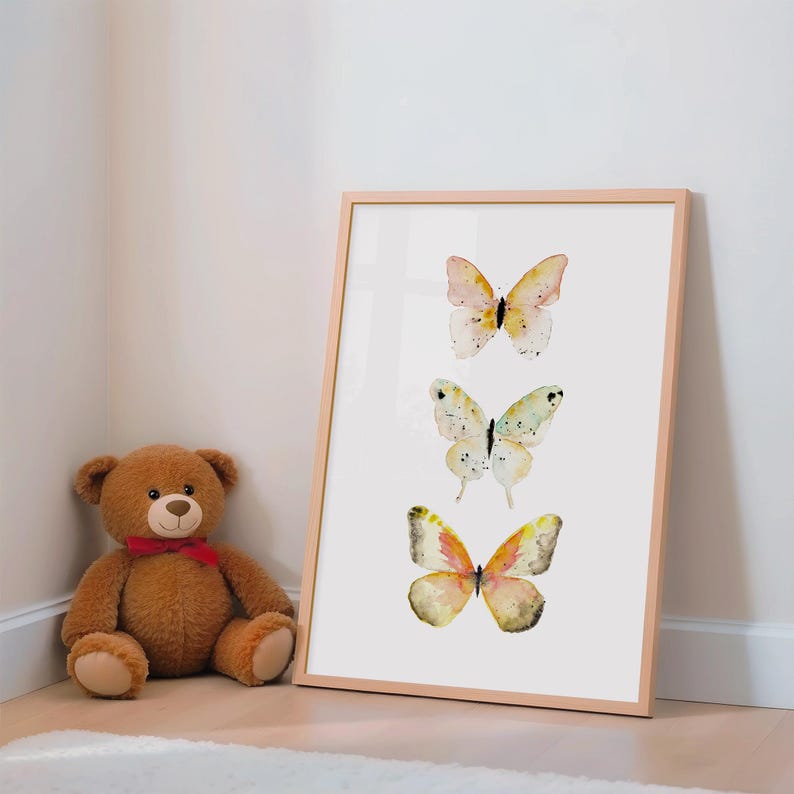 Peut inclure: Une affiche encadr&eacute;e avec trois papillons &agrave; l'aquarelle dans des tons de jaune, rose et vert. Les papillons sont dispos&eacute;s verticalement sur un fond blanc dans un cadre en bois brun clair. Un ours en peluche marron avec un n&oelig;ud rouge est &agrave; proximit&eacute;.
