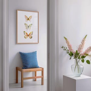 Peut inclure: Une impression encadr&eacute;e avec trois illustrations de papillons &agrave; l'aquarelle dans les tons de jaune, vert et orange. Un coussin bleu &agrave; motifs repose sur un banc en bois sous l'&oelig;uvre d'art. Un vase de fleurs roses est sur un pi&eacute;destal blanc.
