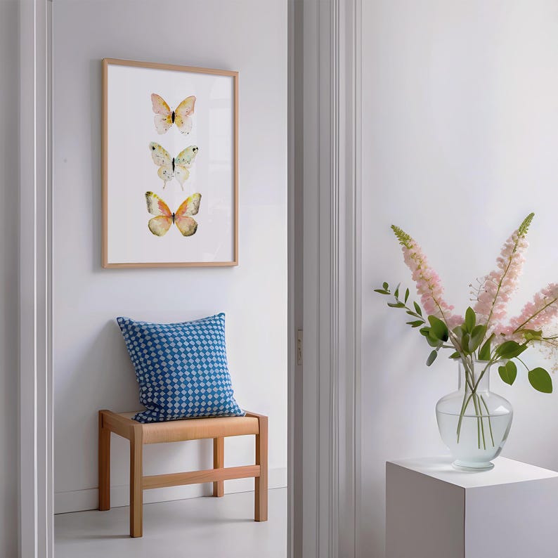 Peut inclure: Une impression encadr&eacute;e avec trois illustrations de papillons &agrave; l'aquarelle dans les tons de jaune, vert et orange. Un coussin bleu &agrave; motifs repose sur un banc en bois sous l'&oelig;uvre d'art. Un vase de fleurs roses est sur un pi&eacute;destal blanc.