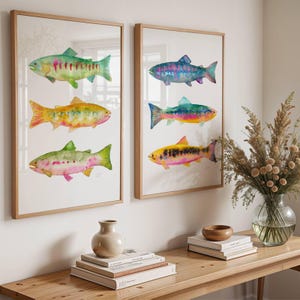May include: Two framed watercolor prints of colorful fish. Each print features three different fish with vibrant hues of green, yellow, pink, and blue. The frames are a light wood color, and the artwork is displayed on a white wall.