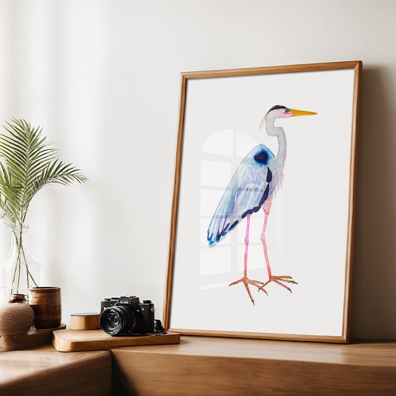 Puede incluir: Una impresi&oacute;n en acuarela enmarcada de una garza. El ave est&aacute; representada con tonos azules, grises y rosas, con un pico amarillo y detalles en negro. La obra de arte se exhibe en un marco de madera, agregando un toque de elegancia natural.
