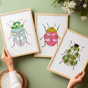 May include: Three framed watercolor paintings of colorful beetles. The beetles are green, pink, and blue with black outlines. The frames are light wood.