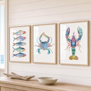 May include: Three framed wall art prints featuring watercolor illustrations of marine life. The left print shows five stacked fish, the center print displays a blue crab, and the right print depicts a colorful lobster, all set against white backgrounds.