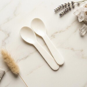 May include: Two off-white spoons are arranged on a marble surface, alongside decorative elements including a sprig of lavender, quartz crystals, and a dried plant. The spoons have a simple, rounded design.