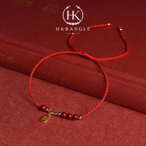 Könnte beinhalten: Rotes Armband mit goldenen Akzenten und einem kleinen goldenen Anhänger. Das Armband hat rote Perlen und ist vor rotem Hintergrund platziert. Das Markenlogo "HK BANGLE" ist oben sichtbar.