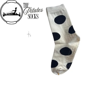 Könnte beinhalten: Eine weiße Pilates-Socke mit großen schwarzen Polka-Dots. Die Socke hat einen strukturierten Grip auf der Sohle und der Text "THE Pilates SOCKS" ist sichtbar.