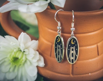 May include: Silver dangle earrings with an oval pendant featuring a bridge design under a crescent moon and stars. The earrings are set against a blurred background of a terracotta pot and white flowers.