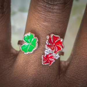 May include: Two open-ended rings with floral designs. One ring features a green leaf design with gold trim. The other ring has a red flower design with clear gemstones. Both rings are silver in color.