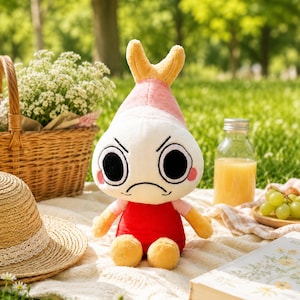 Puede incluir: Un peluche con cuerpo blanco, camisa roja y pies y brazos amarillos. El juguete tiene una cabeza rosa y amarilla con una expresi&oacute;n de mal humor y grandes ojos negros. El juguete est&aacute; sentado en una manta blanca con una cesta de picnic, un sombrero y un libro en el fondo.